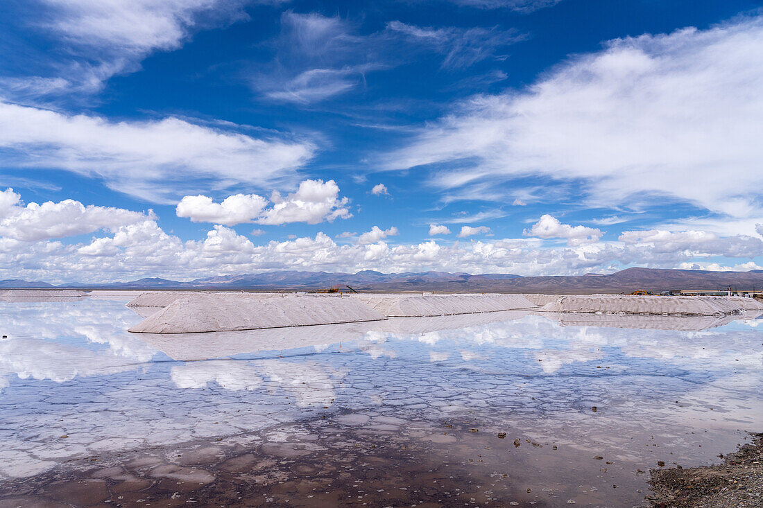 Piles of salt at a salt mining operation on the salt flats of Salinas Grandes in northwest Argentina. Clouds are reflected on a shallow sheet of water.