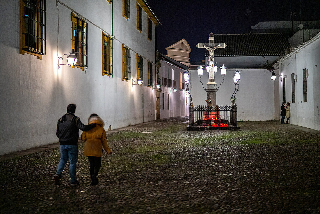 A couple walks through the historic Plaza de Capuchinos in Cordoba, Spain, near the illuminated Cristo de los Faroles, capturing an atmospheric night scene.