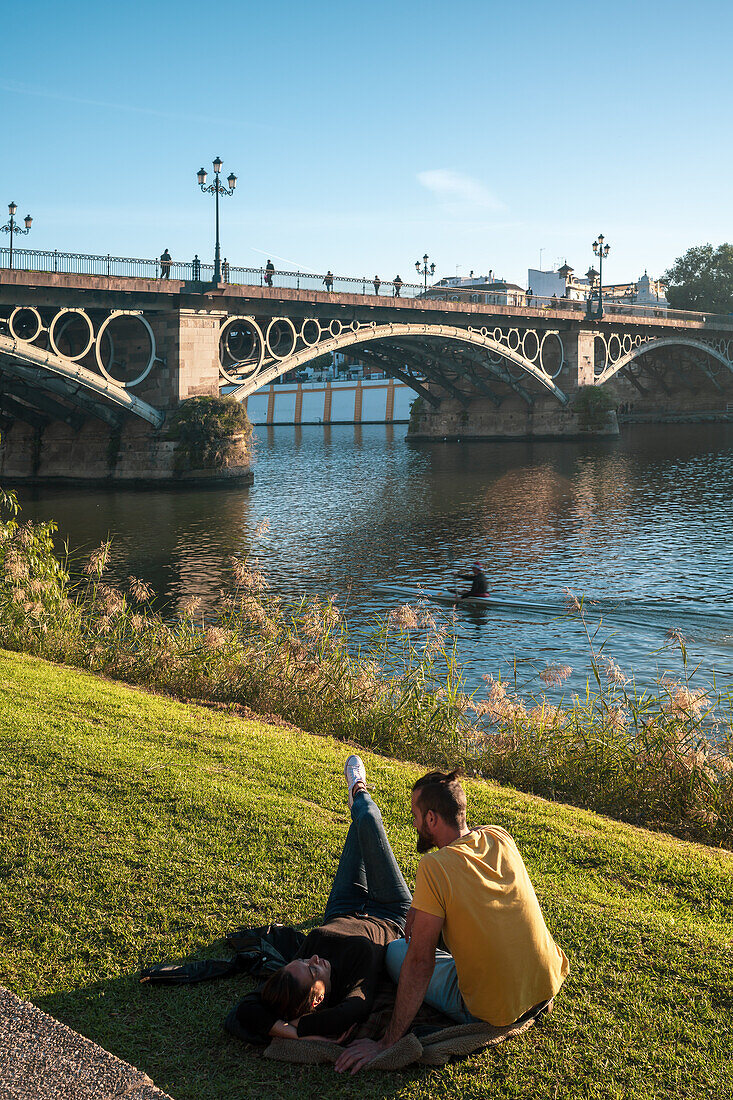 Seville, Spain, Dec 4 2018, A serene moment as a couple relaxes by the riverbank under the iconic Triana Bridge in Seville, Spain. The scene captures harmony and tranquility in a picturesque setting.