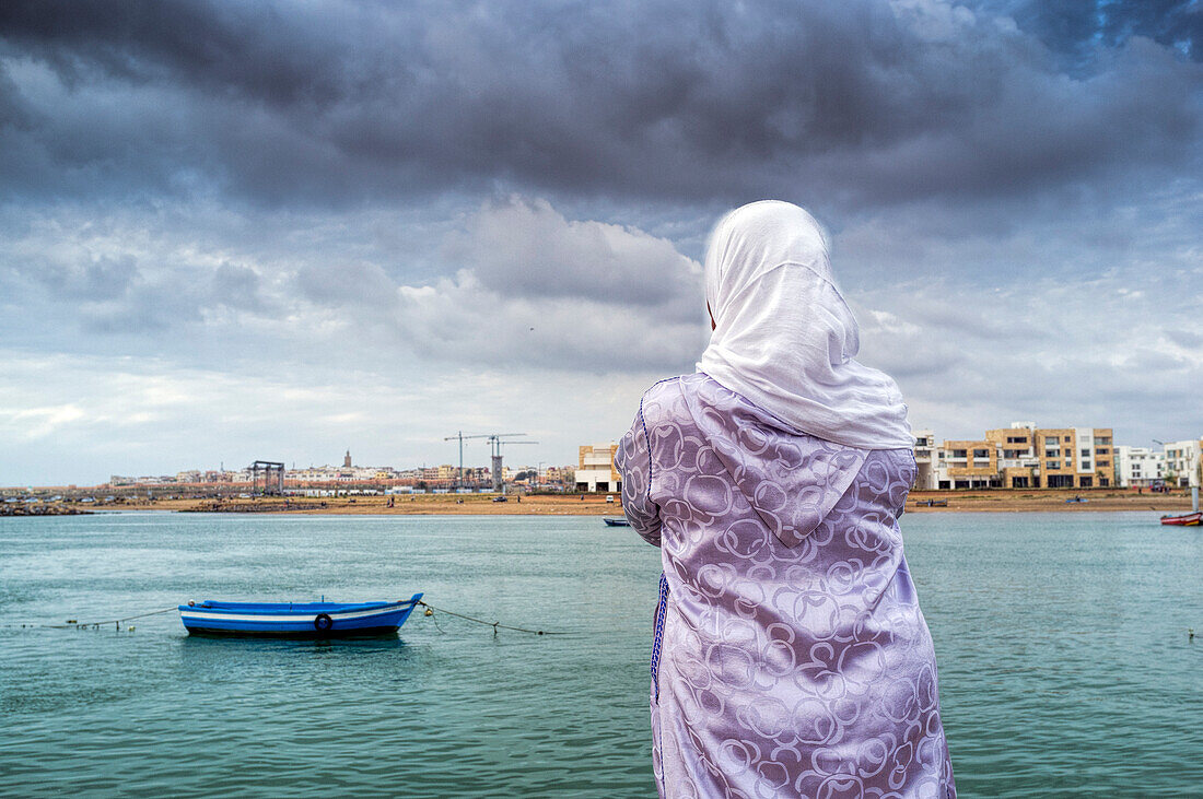 A woman stands by the Bou Regreg River in Rabat, Morocco, admiring the tranquil water and distant cityscape under a cloudy sky.