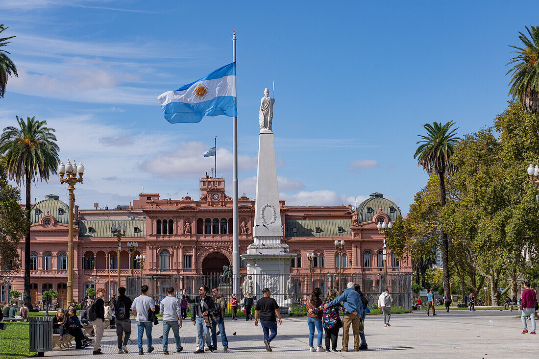 People around an Argentine flag & Pirámide de May or May Pyramid in Plaza de Mayo in front of the Casa Rosada. Buenos Aires, Argentina. The statue represents Liberty.