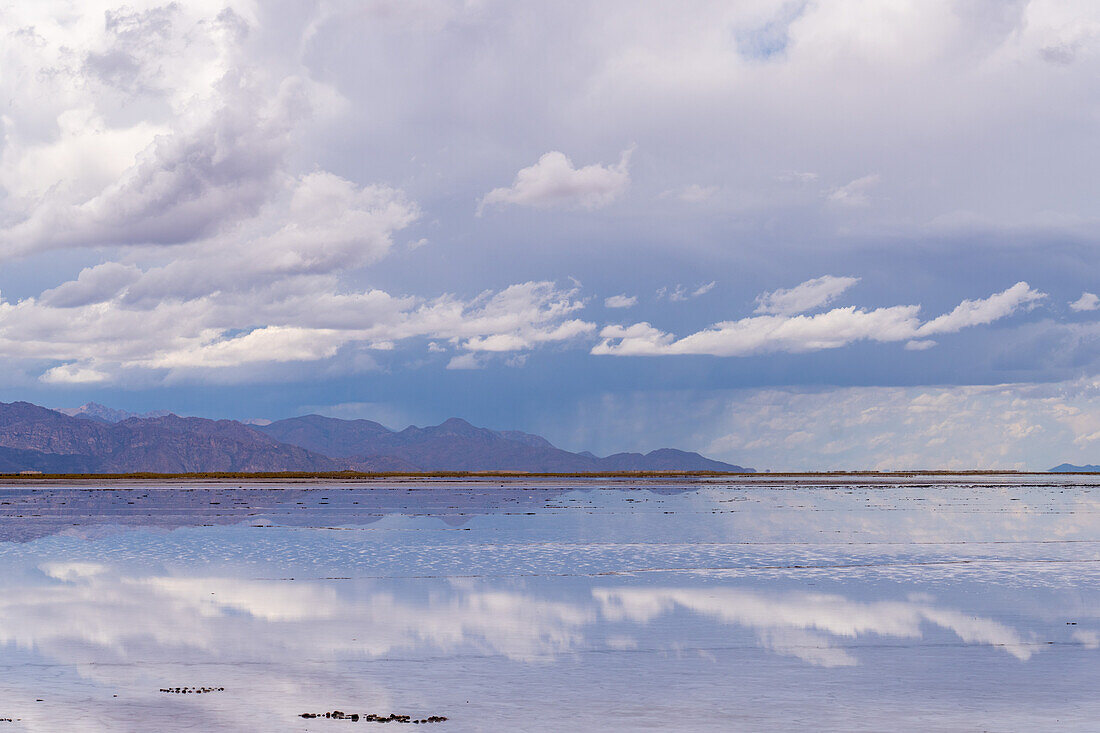 Wolken spiegeln sich auf einer flachen Wasserfläche in den Salinen von Salinas Grandes im Nordwesten Argentiniens, dahinter ein Sturm über den Anden