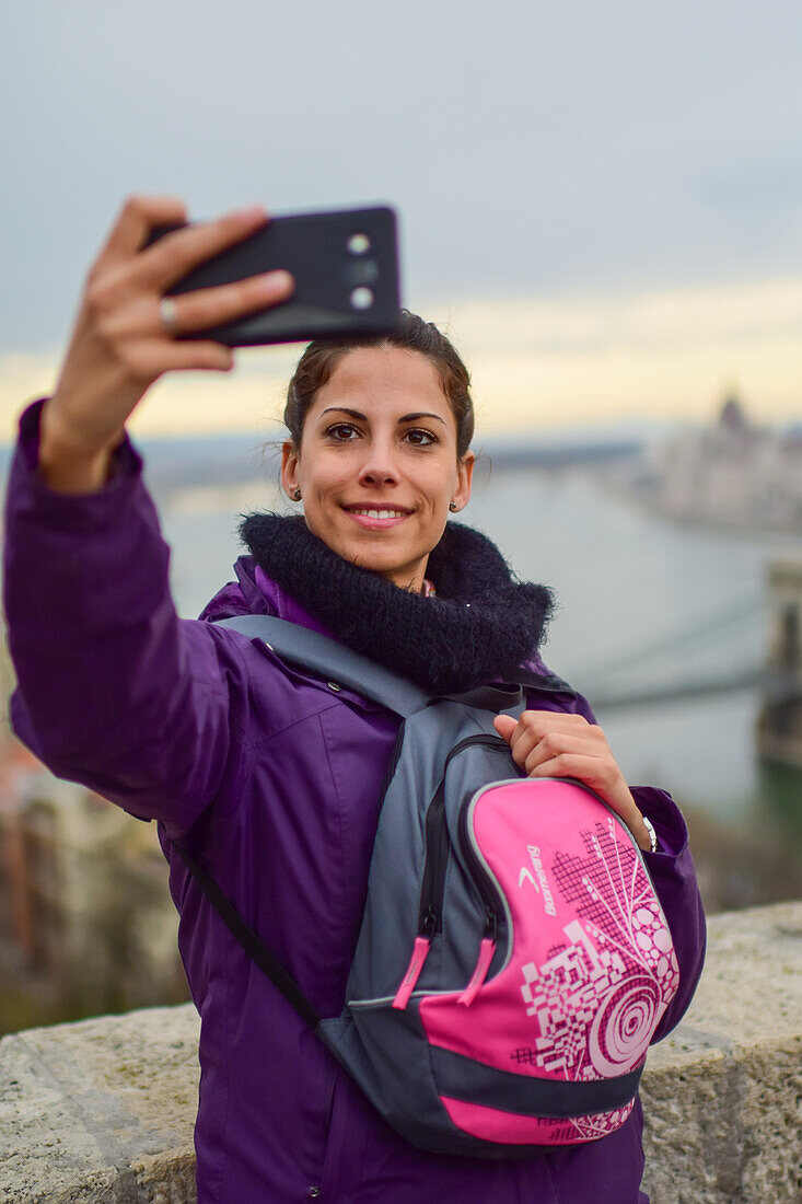 Junge Frau macht ein Selfie mit der Skyline im Hintergrund, einschließlich Parlamentsgebäude und Donau, Budapest, Ungarn, Europa