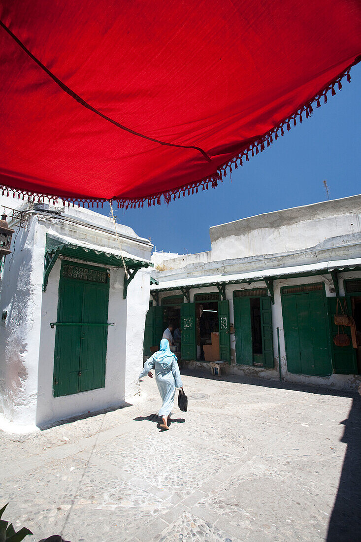 A woman strolls through the historic Medina of Tetouan, surrounded by whitewashed buildings and vibrant green doors under a red canopy.