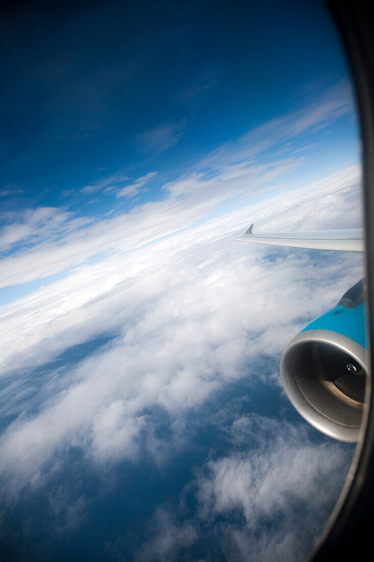 The airplane window reveals a stunning view of clouds below and the wing extending into a clear blue sky during flight.