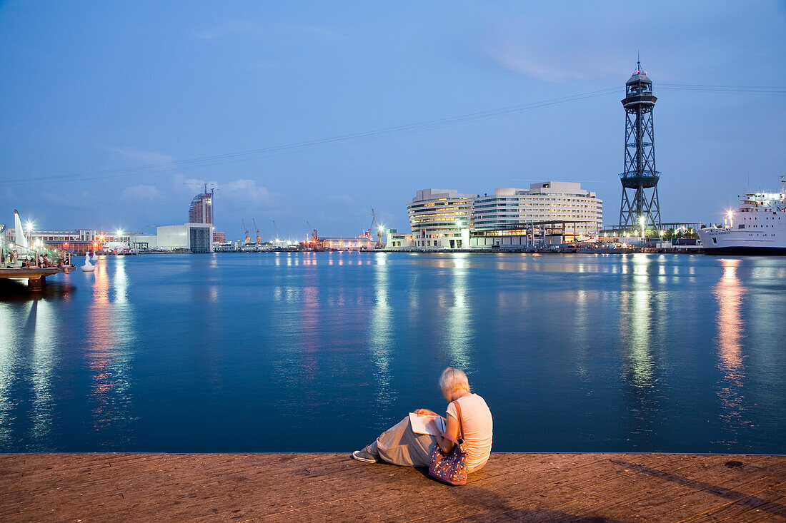 A girl enjoys a quiet moment by the harbor, gazing at the water and city lights as night falls in Barcelona.