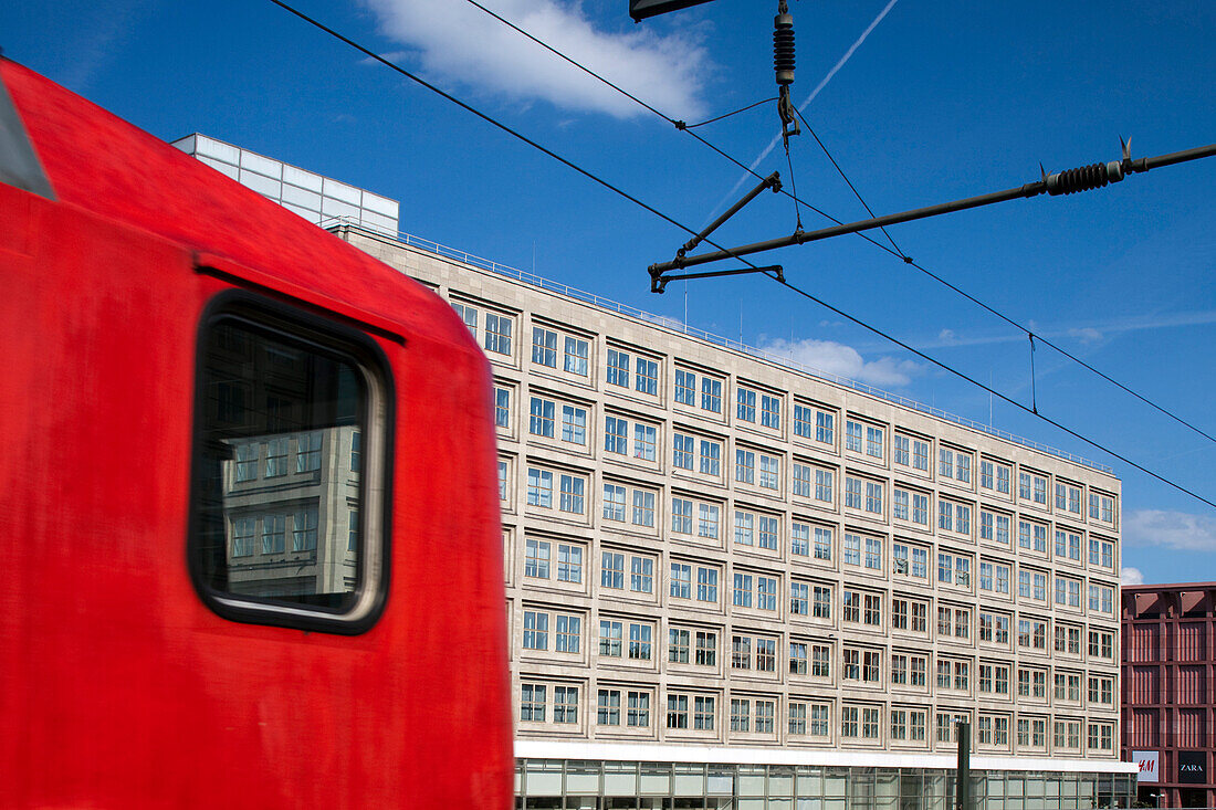 A vibrant red DB Regio train approaches Alexanderplatz station, surrounded by Berlin\'s architectural sights under a bright blue sky.