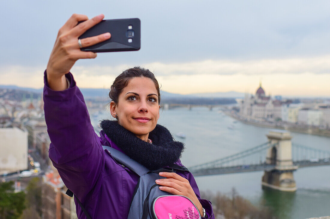 Young woman taking a selfie with skyline behind, including Parliament building and Danube River, Budapest, Hungary, Europe