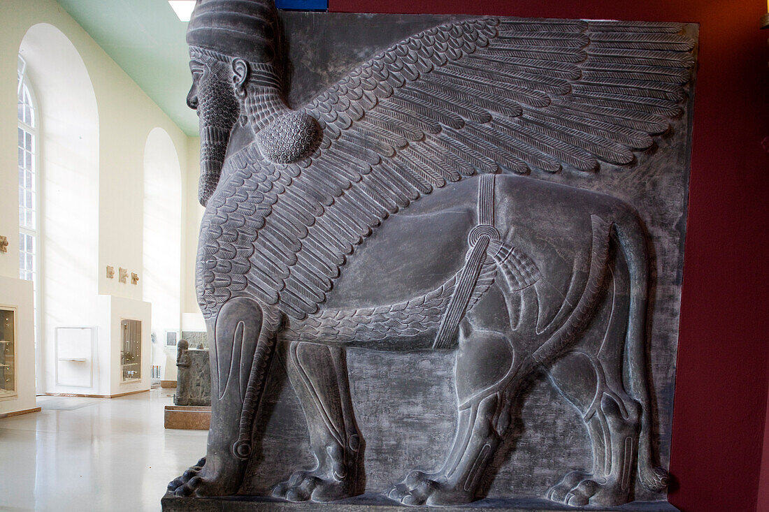 A massive Assyrian winged lion figure with a human head stands in the Pergamon Museum, attracting visitors in Berlin, Germany.