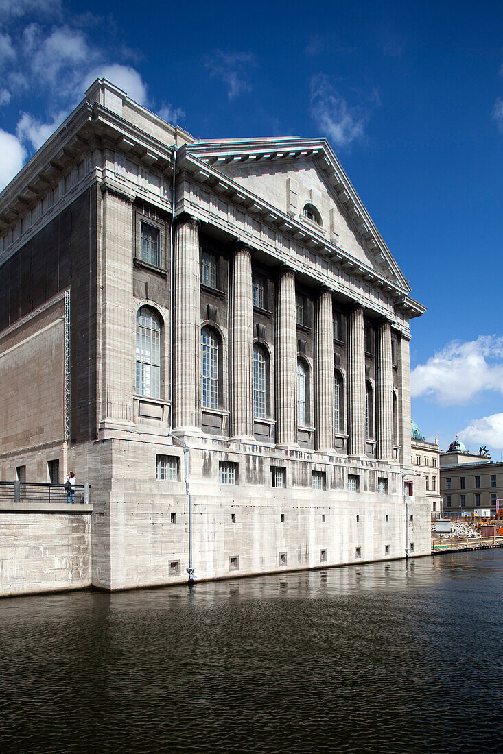 Das Pergamonmuseum, ein Wahrzeichen Berlins, steht majestätisch am Wasser und präsentiert seine beeindruckende Architektur vor einem blauen Himmel