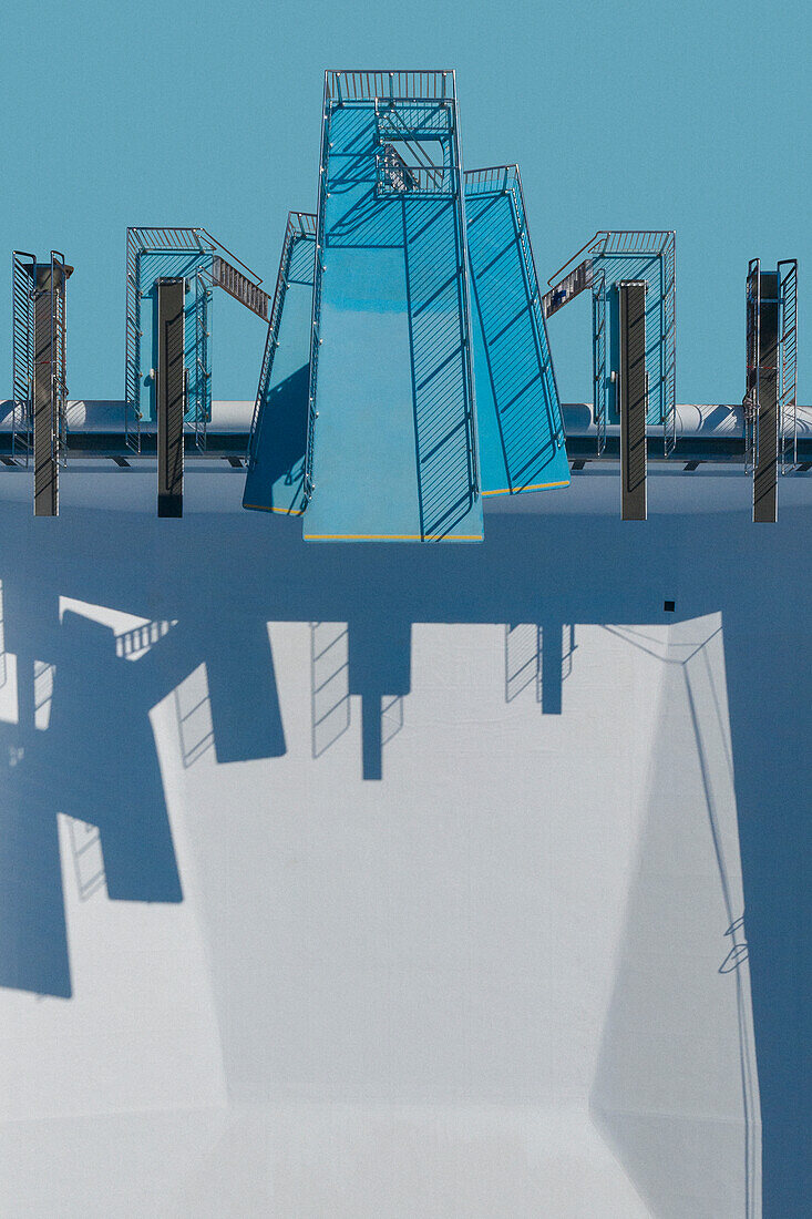 Blick von oben auf Sprungtürme und Sprungbretter, die Schatten über ein sonniges, leeres Schwimmbad werfen