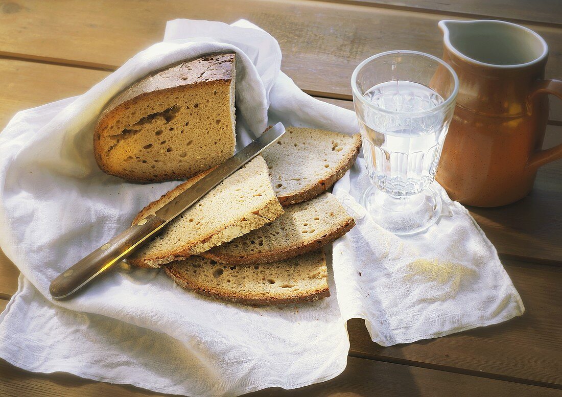 Brot, angeschnitten mit Messer auf Tuch, Wasserglas und Krug