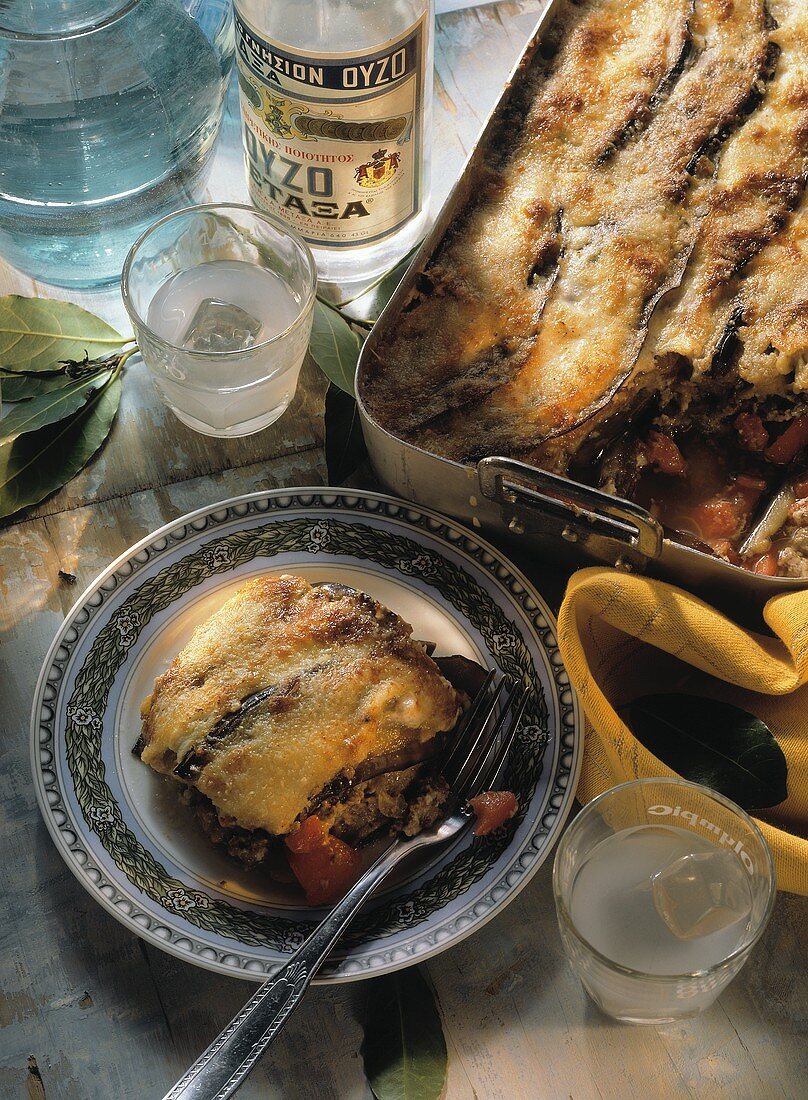 Moussaka in cooking dish and on plate; Ouzo bottle