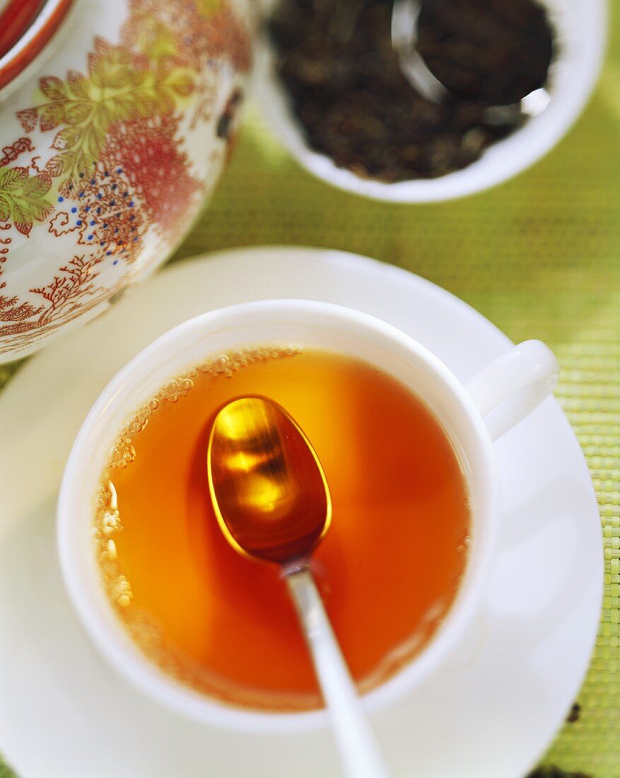 Tasse Tee mit Teelöffel, dahinter Teeblätter