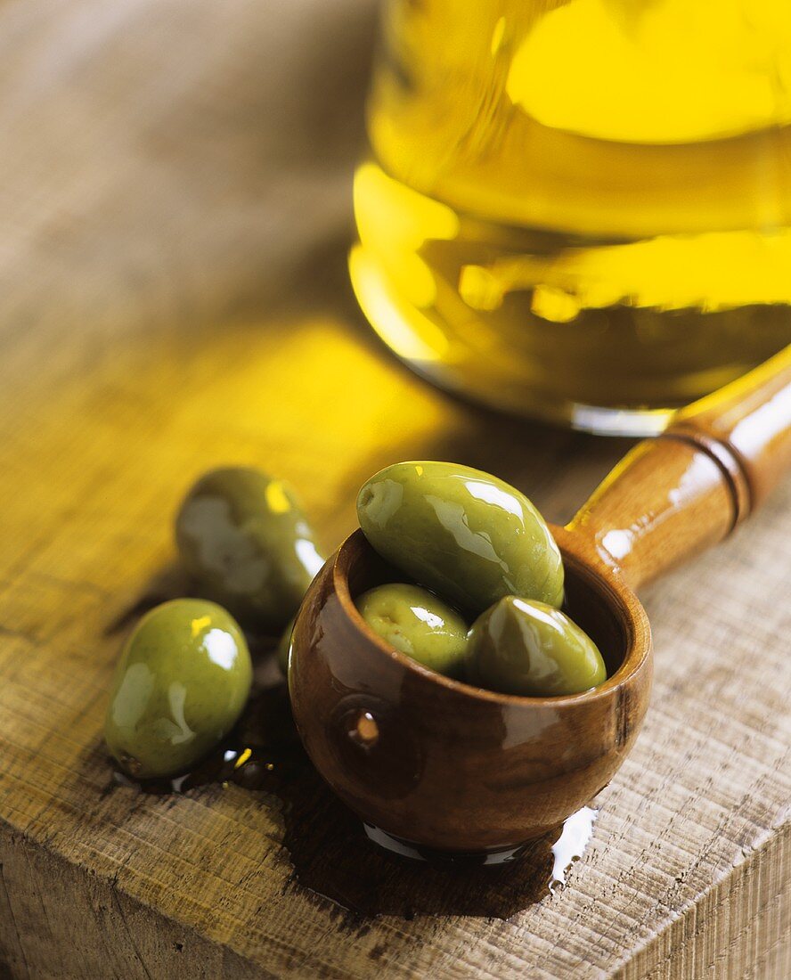 Green olives in olive oil on wooden spoon, bottle of oil behind