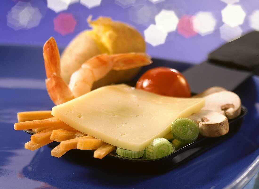 A raclette pan with raw vegetables, shrimps & cheese