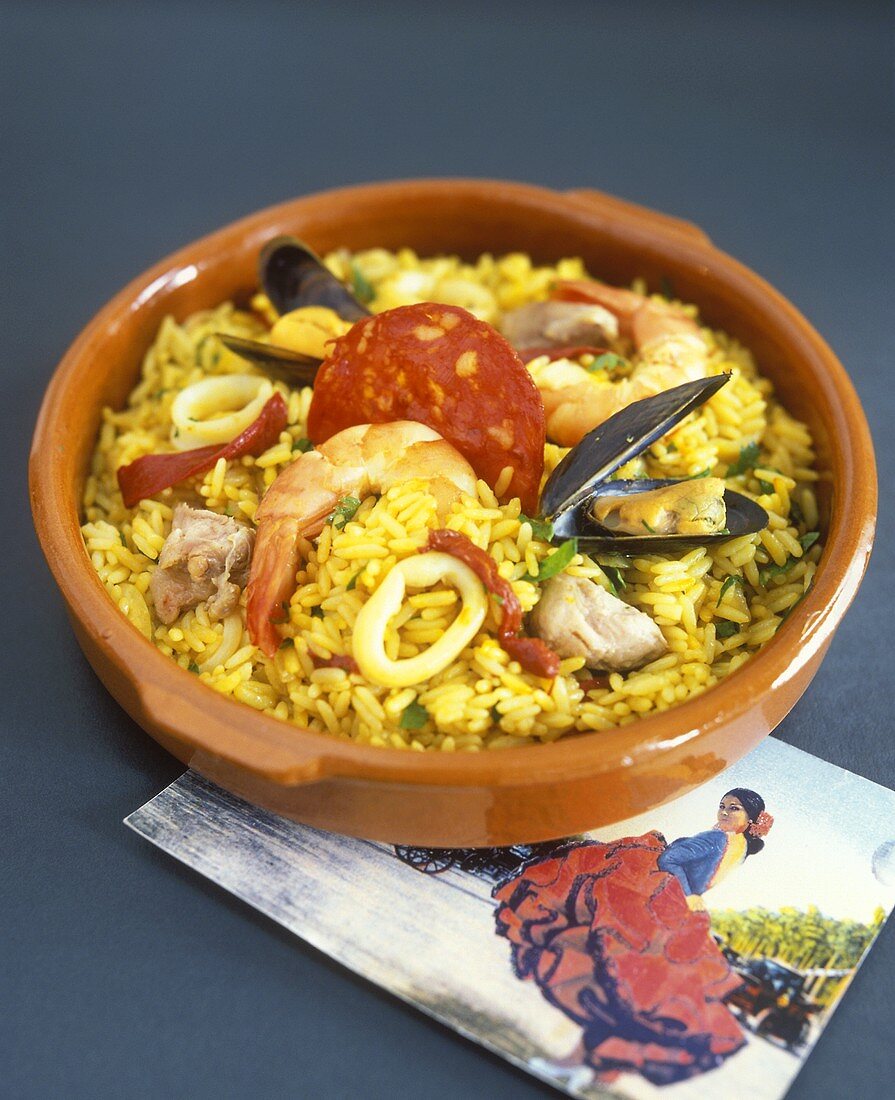 Paella in brown stoneware dish, Spanish postcard beside it