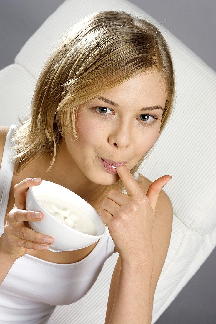 Junge Frau mit Schälchen Joghurt, einen Finger abschleckend