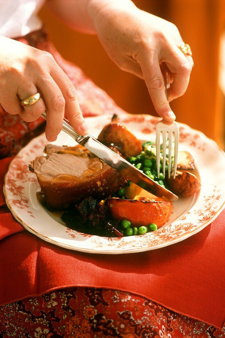 Hands with cutlery over a plate of roast pork with vegetables