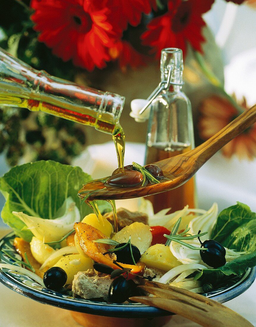Pouring oil on wooden spoon with olives over vegetable salad
