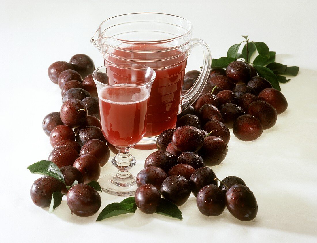 Plum juice in glass and jug surrounded by fresh plums