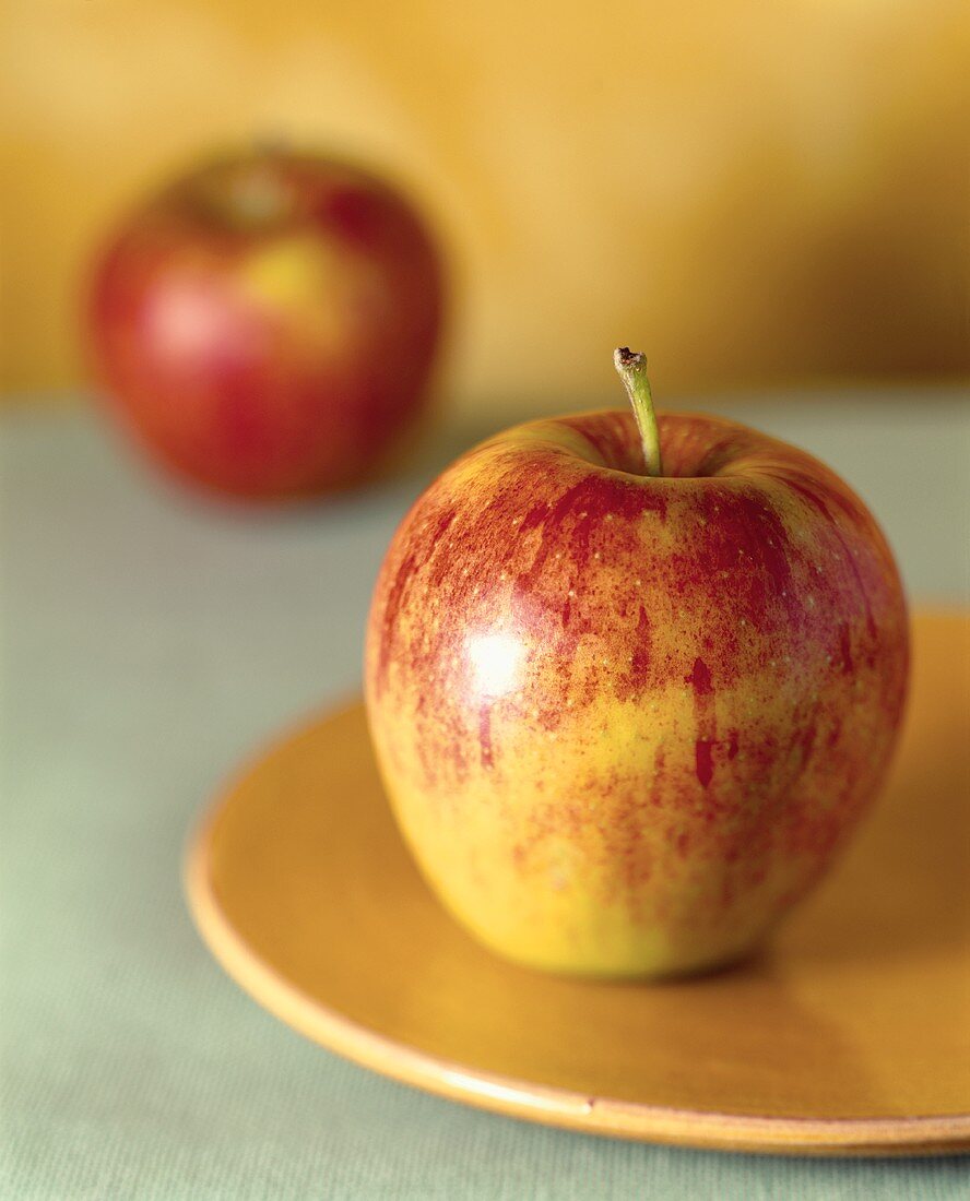 Gelb-roter Apfel auf gelbem Teller vor zweitem Apfel