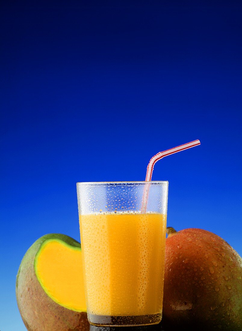 Mango juice in a glass with straw in front of two mangoes