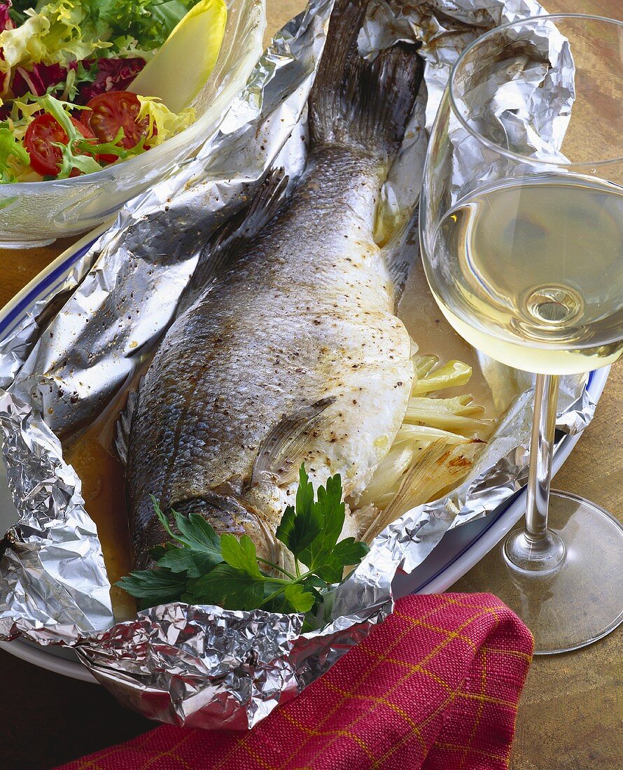 Whole sea perch in foil on platter; white wine glass; salad