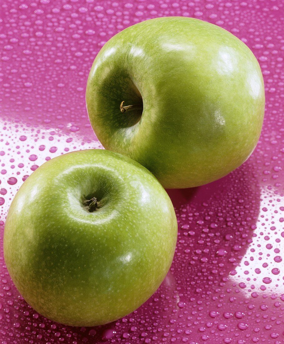 Two Granny Smith apples on a pink background with water drops