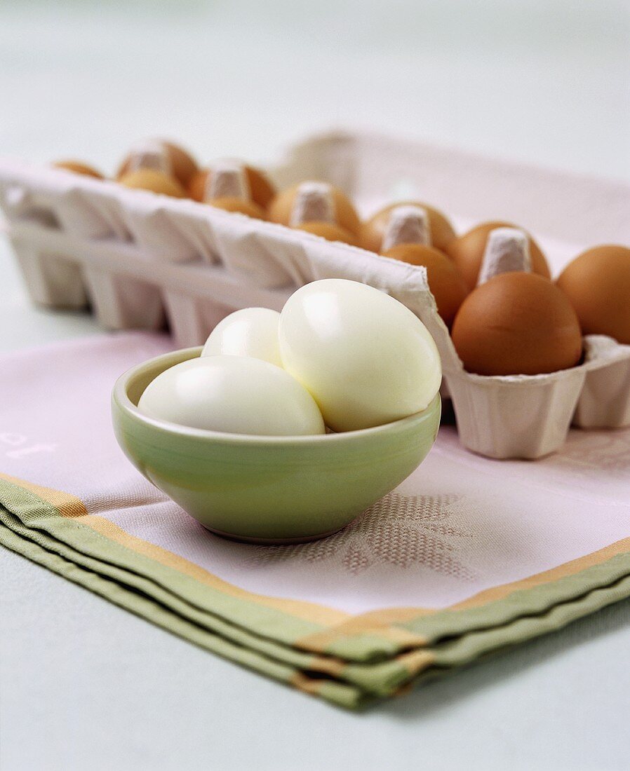 Three shelled boiled eggs, with  eggs in an egg box behind