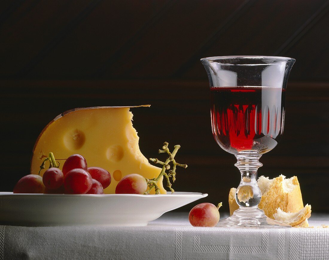 Ein Stück Emmentaler, rote Trauben, Rotwein und Weißbrot