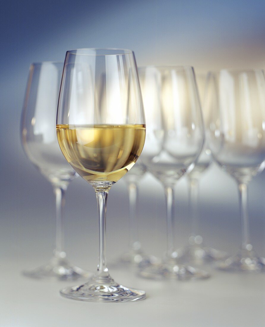 A half filled white wine glass in front of empty glasses