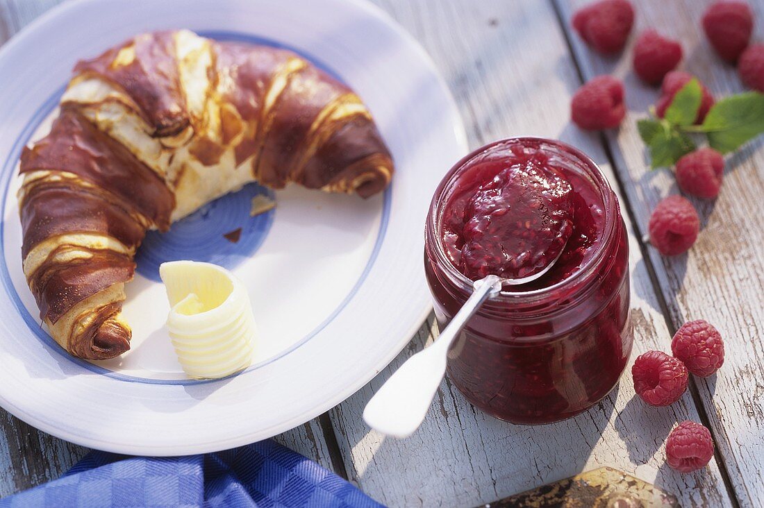 Raspberry jam in jar beside croissant with butter