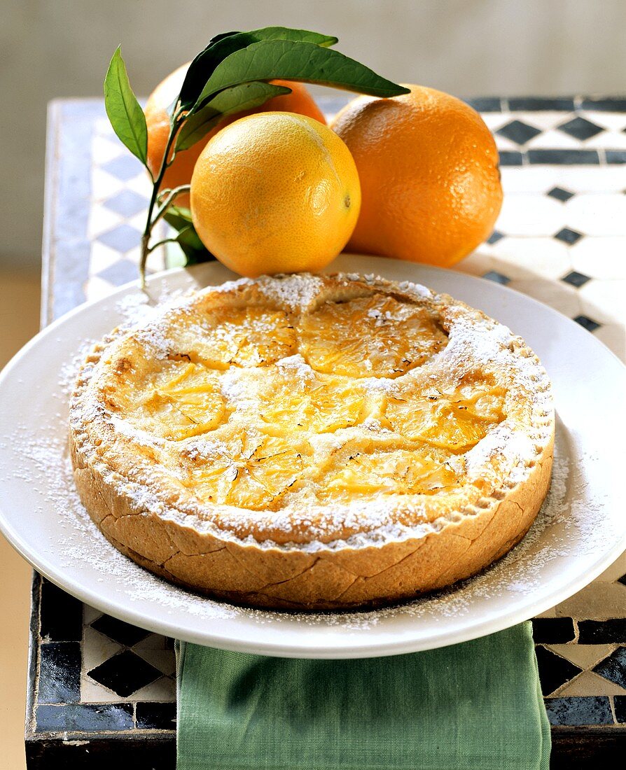 Orange & anise tart with icing sugar on plate; oranges