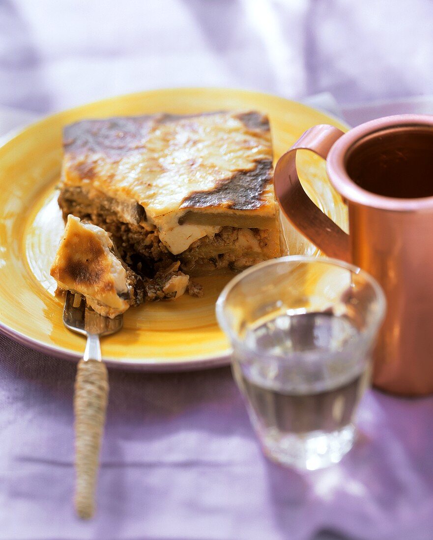 Moussaka on yellow plate and fork; wine; copper jug