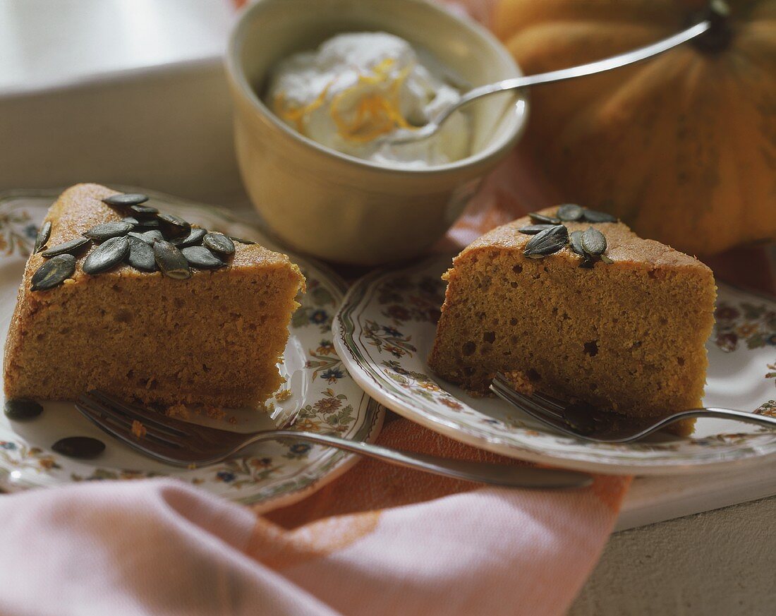 Two pieces of pumpkin cake with pumpkin seeds; cream in bowl