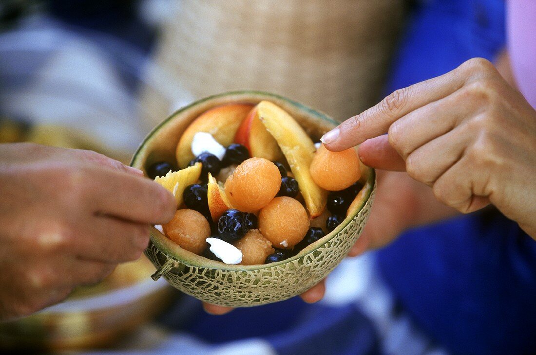 Hands reaching for pieces of fruit salad in halved melon