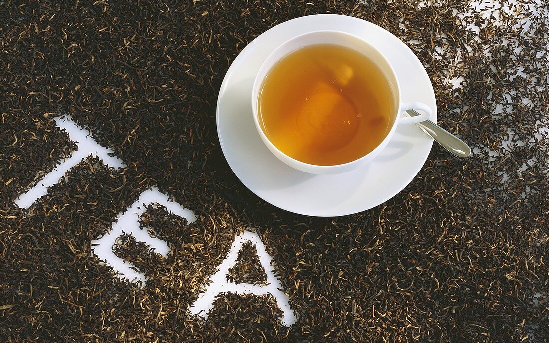 Tasse Schwarztee, umgeben von Teeblättern mit der Schrift TEA