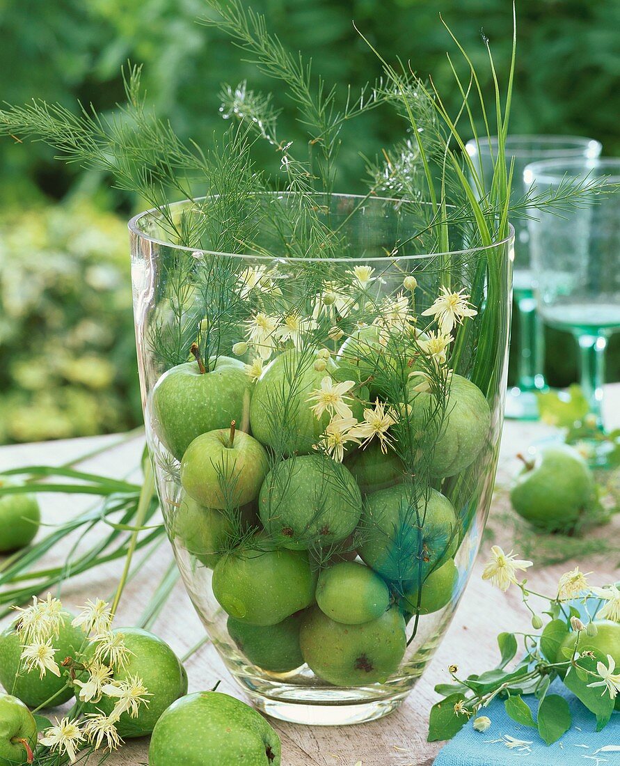 Grüne Äpfel, Zierspargel und Waldrebe in grosser Glasvase
