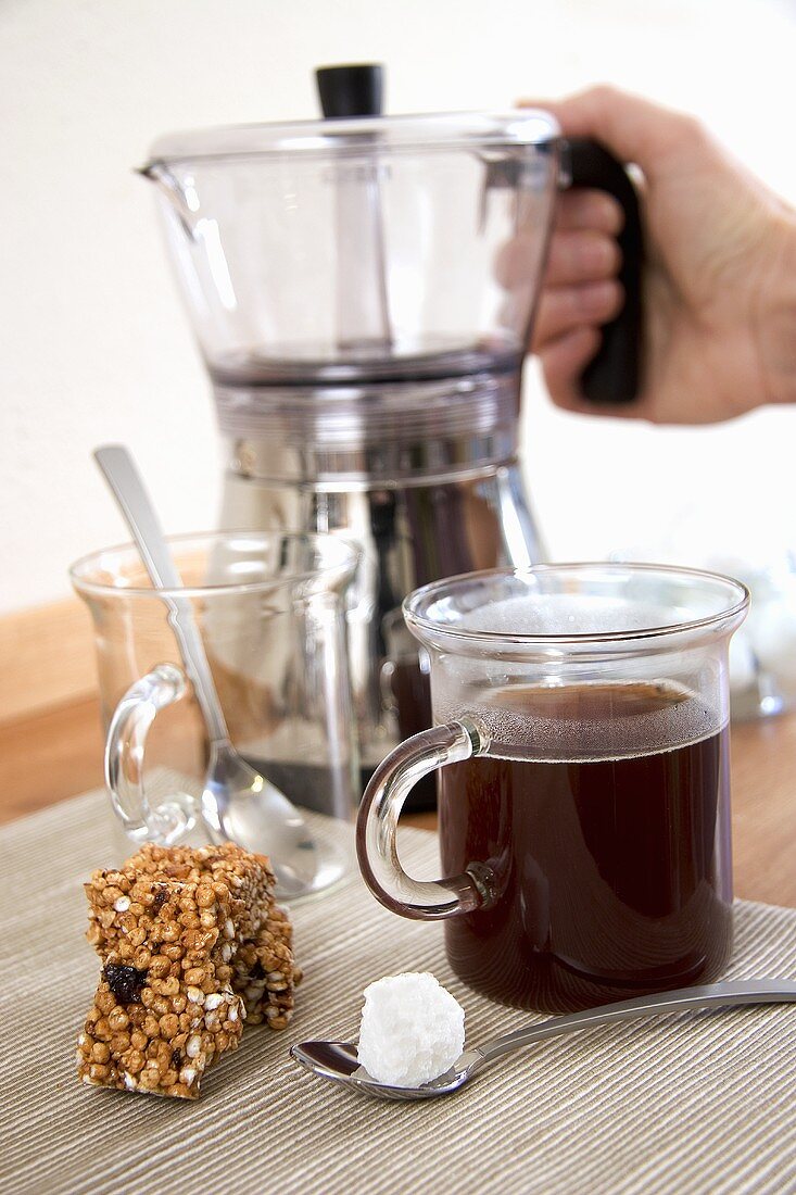 Coffee in glass mug, sugar cube, muesli bar, coffee maker