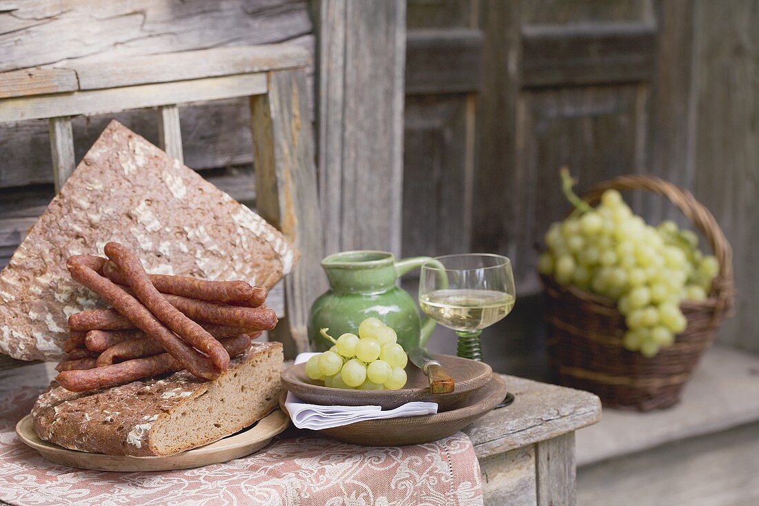Trauben, Brot, Wurst und Wein auf Holzbank vor Bauernhaus
