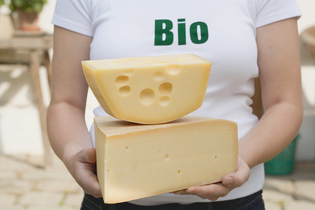 Frau hält zwei grosse Stücke Bio-Käse vor einem Bauernhaus