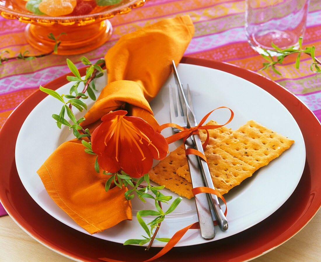 Place-setting with orange napkin, fire lily & crispbread