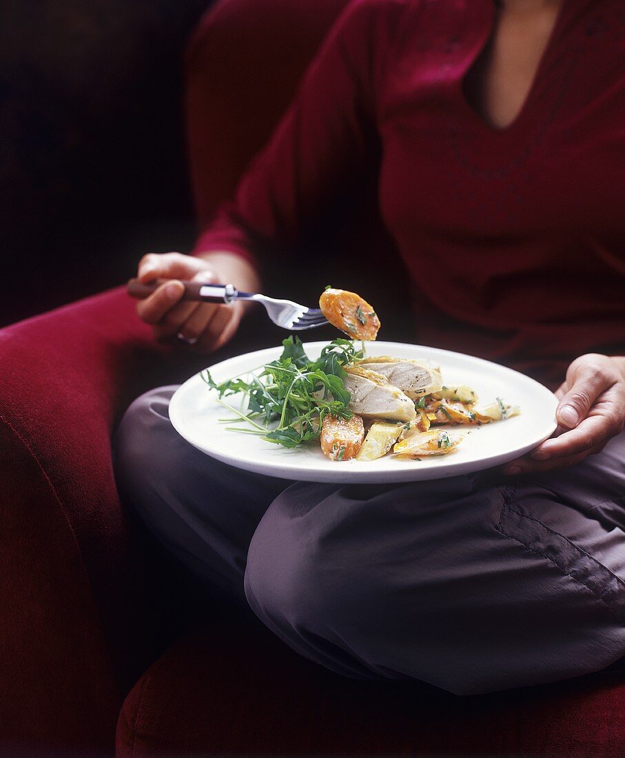 Woman eating chicken breast with vegetables on sofa