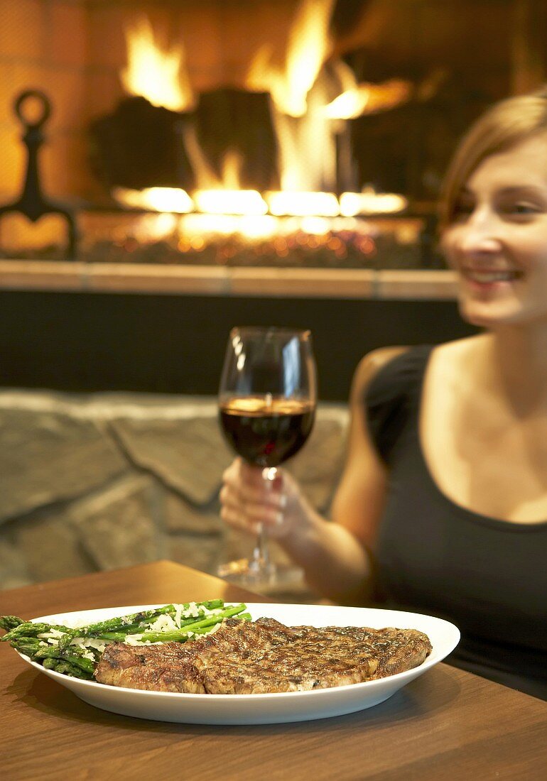 Young woman with rib steak & red wine in front of open fire