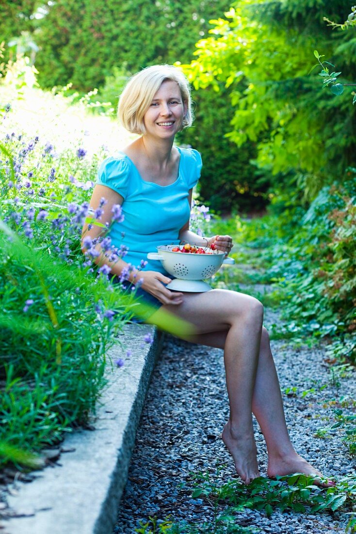 Frau hält Sieb mit Kirschen und sitzt auf Mauer im Garten