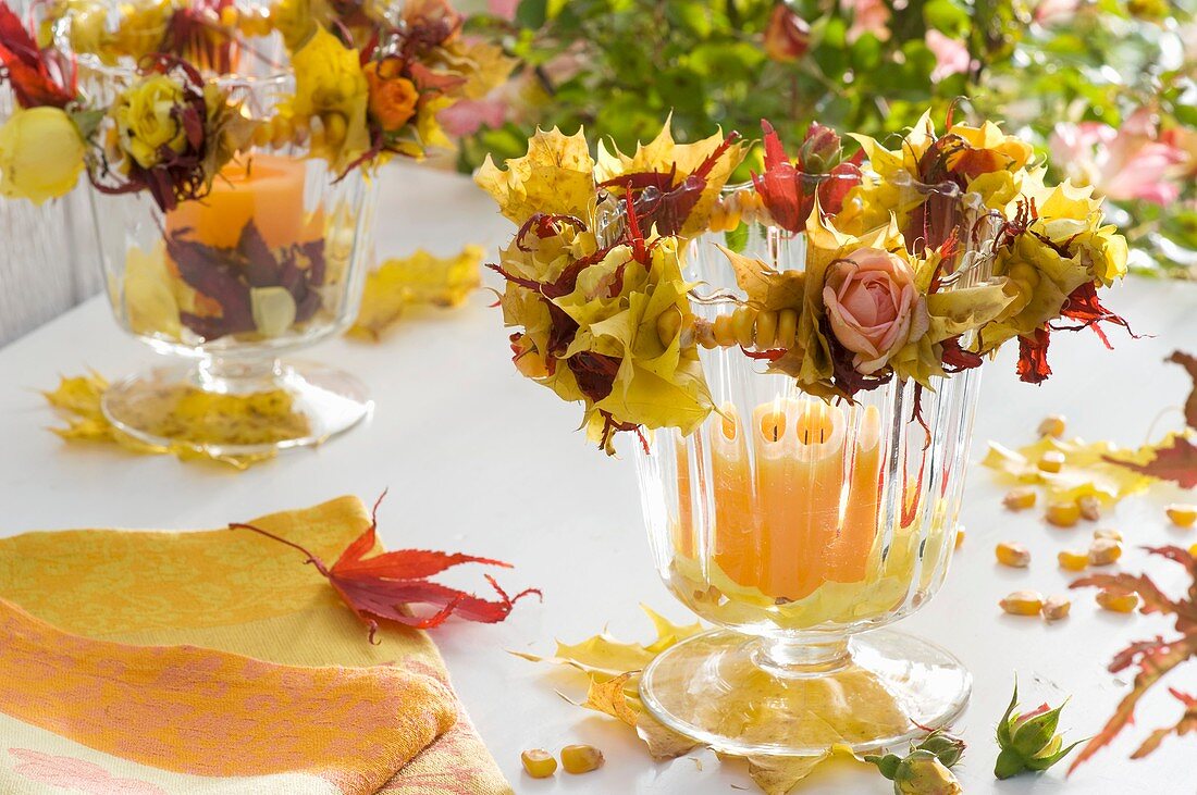 Candles in glasses with wreaths of roses, autumn leaves & corn kernels