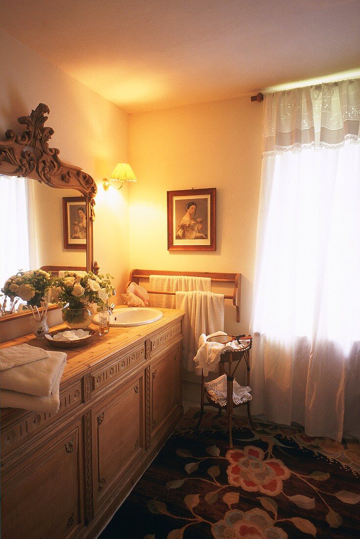 Traditionelles Badezimmer mit Holzkommode als Waschtisch, großem, gerahmten Spiegel, Teppich mit floralem Muster und blickdichtem Fenstervorhang mit Spitzenbesatz