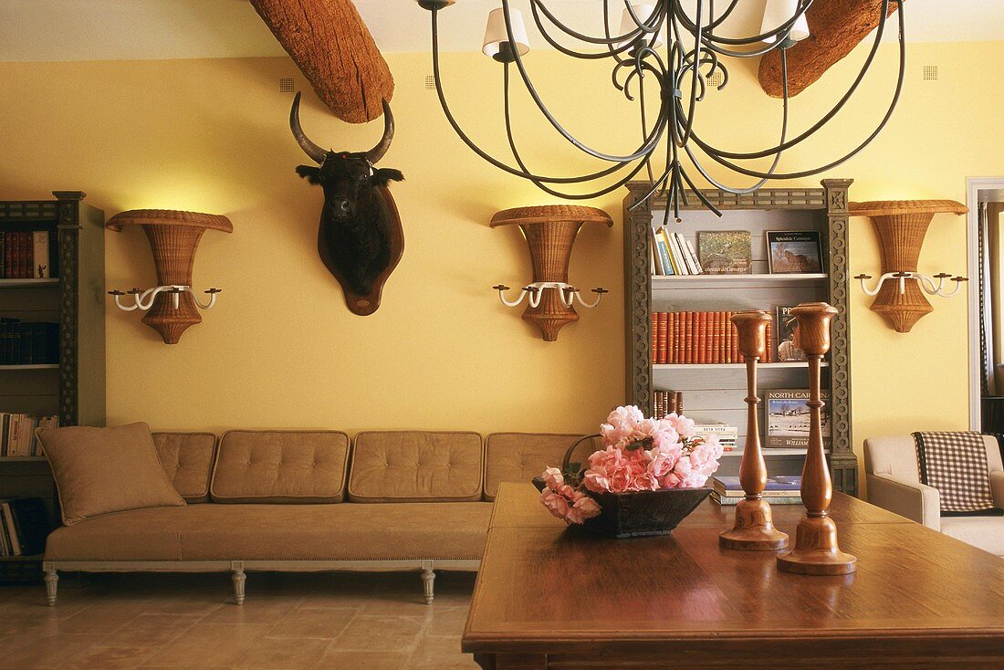 Living room with massive ceiling beams, bulls head, vase-shaped wicker wall lamps and delicate iron chandelier above wooden table with flower arrangement