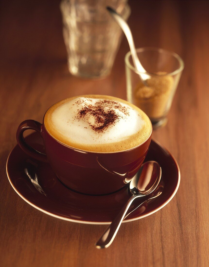 Cappuccino in brown cup and saucer, sugar in glass behind